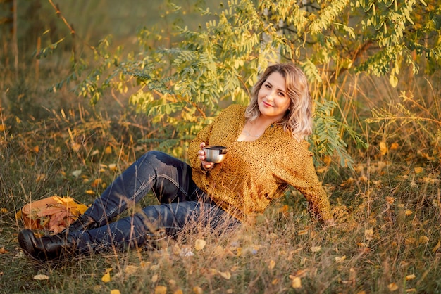 A jovem linda mulher plussize em uma camisa laranja e jeans está sentada em um parque de outono bebendo chá e relaxando em um piquenique