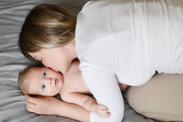 Foto a jovem mãe alegre brinca com seu bebê deitado na cama, sorri, beija a bochecha, desfruta da maternidade
