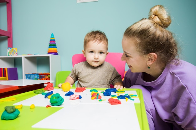 A jovem mãe com seu filho pequeno na sala de jogos esculpir plasticina O menino está sentado à mesa e brincando com plasticina e uma mãe feliz está olhando para ele