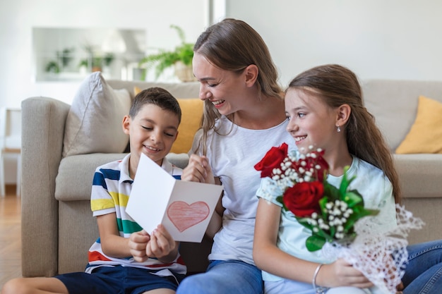 A jovem mãe com um buquê de rosas ri, abraçando o filho, e uma menina fervorosa com um cartão e rosas parabeniza a mãe durante a celebração do feriado em casa. dia das mães