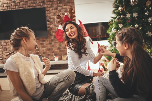 A jovem mãe e suas filhas estão sentadas no chão ao lado da árvore de Natal, rindo e gostam de se olhar felizes.