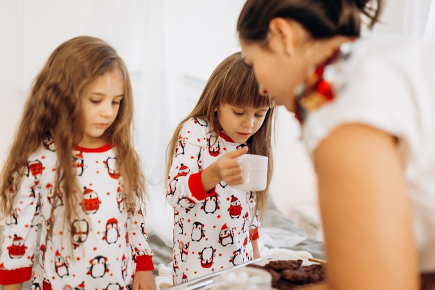 A jovem mãe está trazendo chocolate quente com marshmallows e biscoitos para as filhas de pijama, sentadas na cama em um quarto aconchegante e cheio de luz.