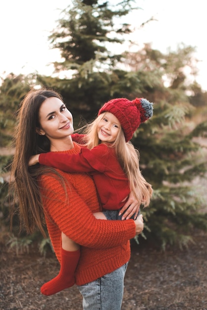 A jovem mãe passa tempo com a filha garota usa suéter de malha vermelha e chapéu sobre a árvore ao ar livre Maternidade Temporada de outono Felicidade