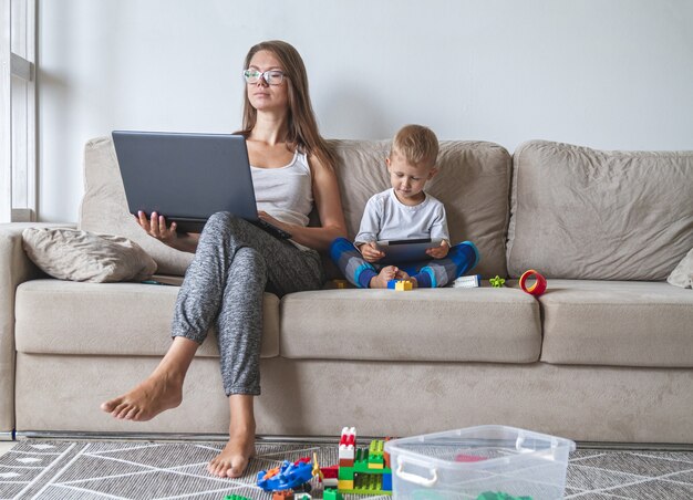 A jovem mãe trabalha em casa sentada no sofá com o laptop enquanto o filho brinca com o tablet