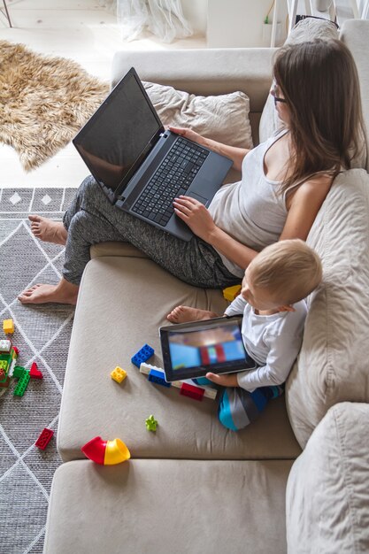 A jovem mãe trabalha em casa sentada no sofá com o laptop enquanto o filho brinca com o tablet