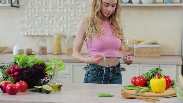 A jovem mede a cintura na cozinha perto da mesa com legumes e frutas Conceito de alimentação saudável e dieta