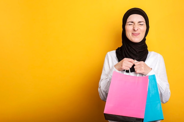 A jovem muçulmana em hijab segura sacolas de compras fechando os olhos e se alegra em um banner de fundo amarelo
