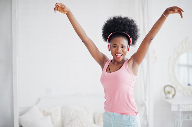 A jovem mulher afro-americana na camisola interioa cor-de-rosa que dança e ouve música em fones de ouvido em seu quarto branco.