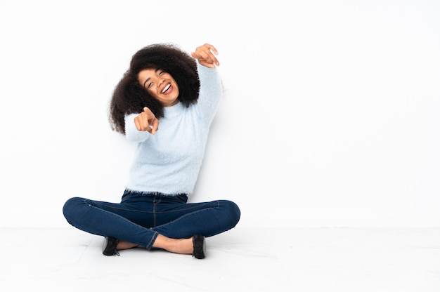 A jovem mulher afro-americana sentada no chão aponta o dedo para você enquanto sorrindo