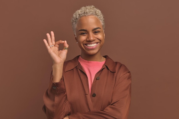 A jovem mulher afro-americana sorridente faz um gesto ok no estúdio marrom