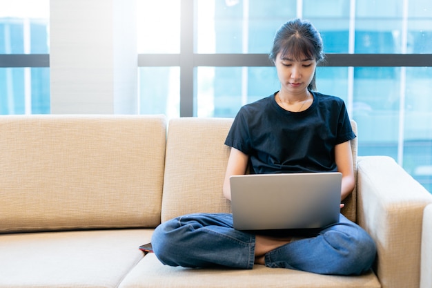 A jovem mulher asiática que trabalha com o portátil da casa, freelance usando o portátil que trabalha pelo Internet, trabalha do conceito home.