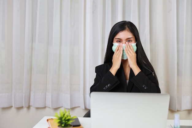 A jovem mulher asiática waring protege a máscara sentado no escritório e trabalhando no computador, laptop e smartphone.