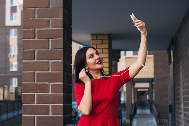 A jovem mulher bonita com batom vermelho no vestido vermelho usa o telefone para tomar o selfie