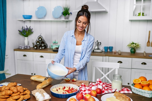 Foto a jovem mulher bonita derrama a massa para a torta de maçã na cozinha branca.