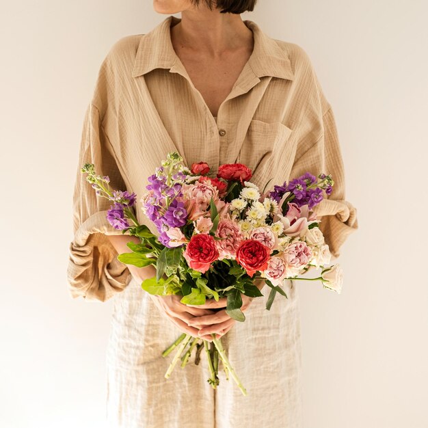 A jovem mulher bonita na camisa de musselina bege segura o buquê de flores rosas coloridas contra a parede branca Bela composição floral de celebração do feriado