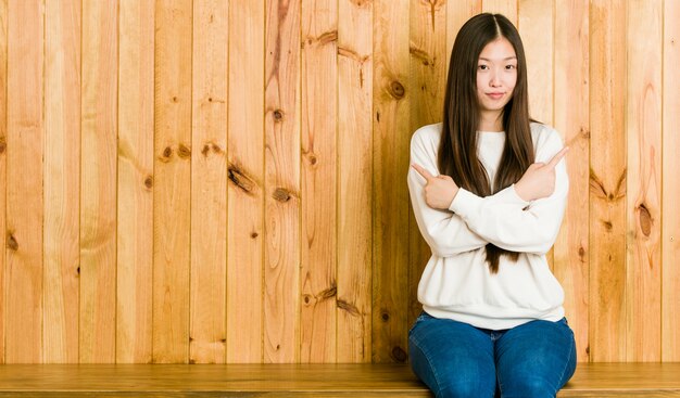 A jovem mulher chinesa que senta-se em um lugar de madeira aponta lateralmente, está tentando escolher entre duas opções.