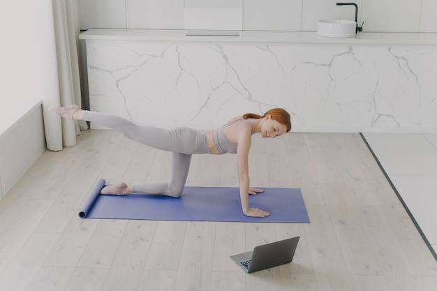 A jovem mulher europeia feliz está fazendo exercícios de prancha e olhando para o laptop workout online em casa