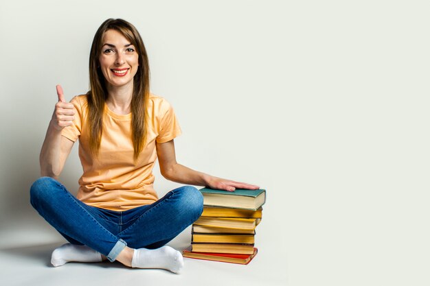 A jovem mulher sorri e faz um gesto com a mão segura a mão dela sobre uma pilha de livros sentada no chão em um espaço claro. Conceito de educação.