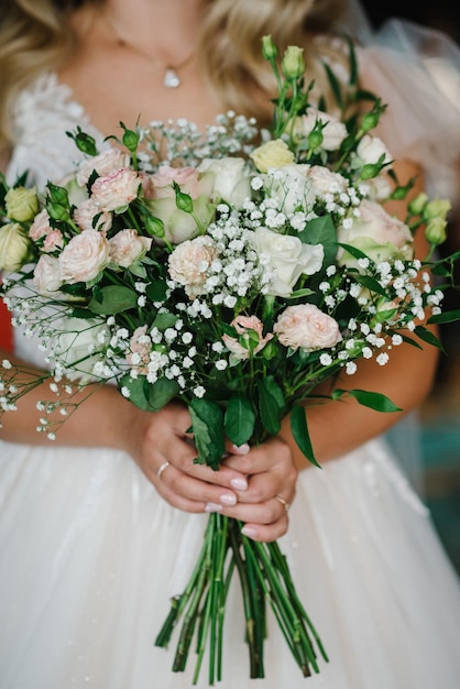 A jovem noiva de vestido elegante está de pé e segurando o buquê de flores cor de rosa e verdes na natureza