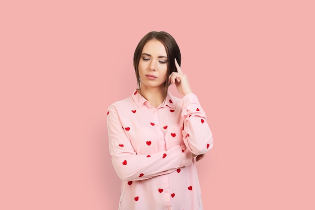 Foto a jovem pensou segurando um dedo na cera isolado em fundo rosa em uma camisa rosa com corações vermelhos