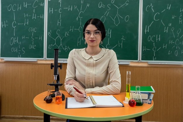 A jovem professora de química senta-se em uma mesa com reação química do microscópio