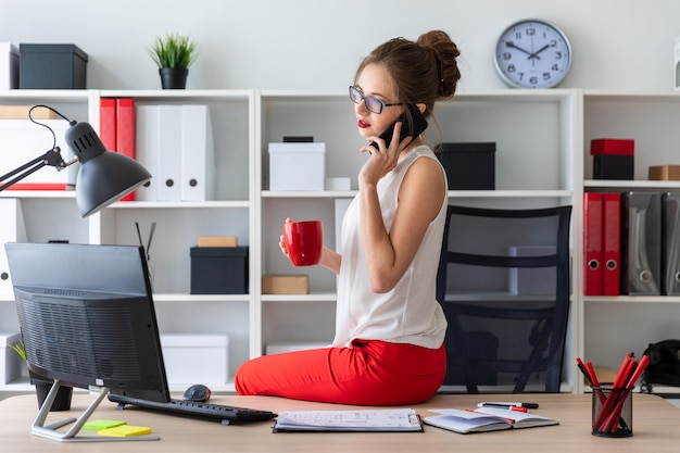 A jovem se sentou na mesa do escritório, segurando um copo vermelho na mão e falando ao telefone.