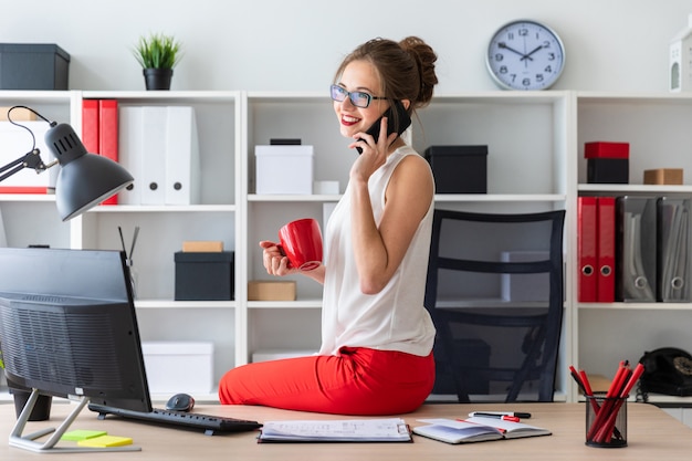 A jovem se sentou na mesa do escritório, segurando um copo vermelho na mão e falando ao telefone.