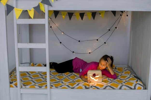 Foto a linda garota está pronta para dormir na cama. noite à noite. menina deitada na cama em beliche decorada com estilo e olhando para o abajur de madeira.