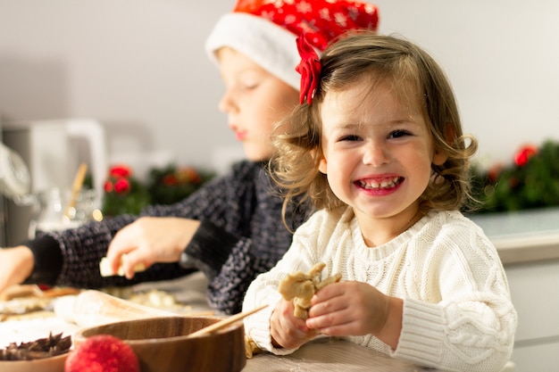 A linda menina 2-4 com um laço vermelho e o menino 7-10 com um boné de Natal fazem biscoitos de gengibre na cozinha de ano novo.