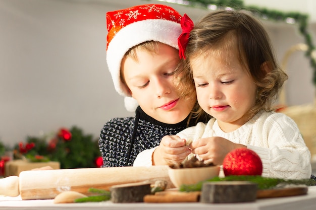 A linda menina 2-4 com um laço vermelho e o menino 7-10 com um boné de Natal fazem biscoitos de gengibre na cozinha de ano novo.