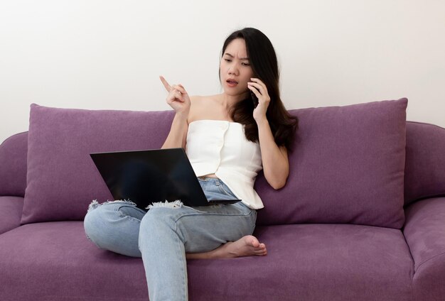 A linda mulher asiática está na sala de estar e sentada no sofá com o laptop está no smartphone para conversar e discutir sobre um negócio ou trabalho