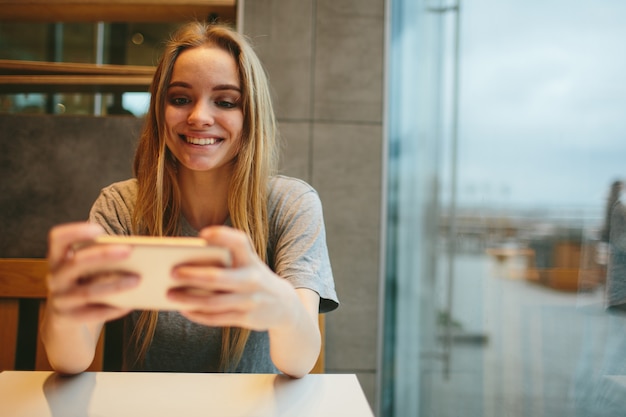 A loira usa o telefone. Menina e smartphone. Uma mulher está sentada em um café com um celular