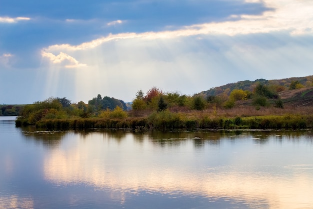 A luz do sol penetra pelas nuvens sobre o rio e a floresta ao pôr do sol. As nuvens são refletidas na água do rio.