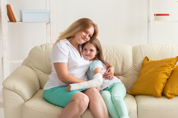 A mãe abraça a sua filha com um gesso no pulso ou braço quebrado sorrindo. Doenças infantis, perspectiva positiva e recuperação.