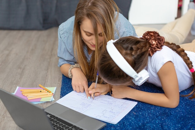 Foto a mãe ajuda a filha a fazer o dever de casa na frente do monitor de um laptop deitada no chão