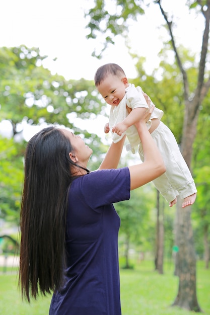 A mãe asiática joga acima o bebê infantil feliz nos braços no jardim.