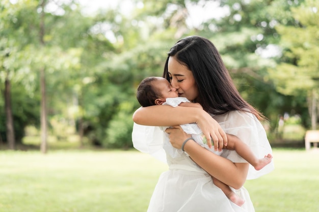 A mãe bonita nova asiática que guarda seu recém-nascido está dormindo e sente-se com amor e tocando-se delicadamente então sentando-se na grama verde no parque