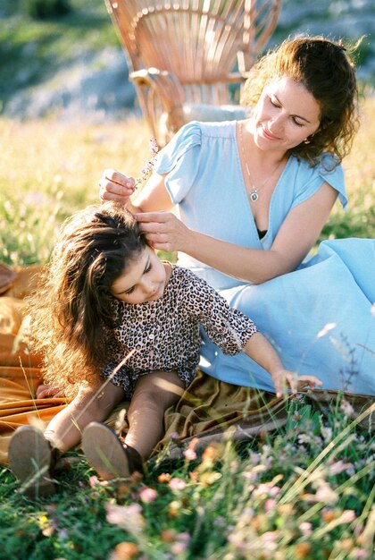 Foto a mãe decora o cabelo de uma menina com flores silvestres sentada em uma colcha de cama em um prado ensolarado