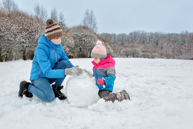 Foto a mãe e a filha brincam ao ar livre com máscaras médicas e fazem um boneco de neve com bolas de neve