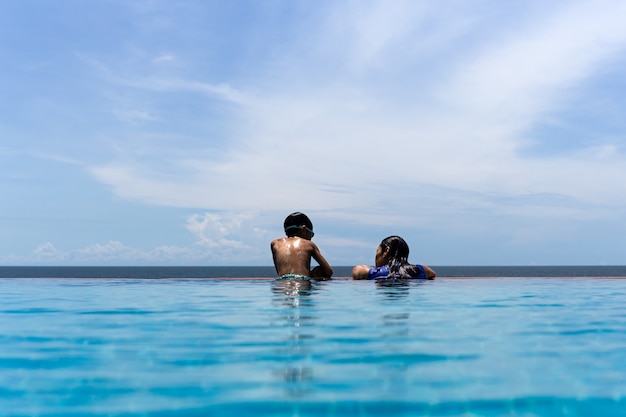 A mãe e o filho novo relaxam na piscina que olha sobre o oceano em férias de verão.