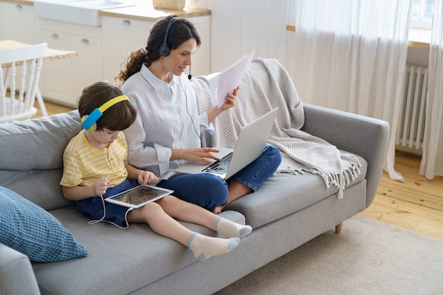 A mãe freelancer sentada no sofá no escritório em casa durante o trabalho de bloqueio no laptop, criança brincando de tablet