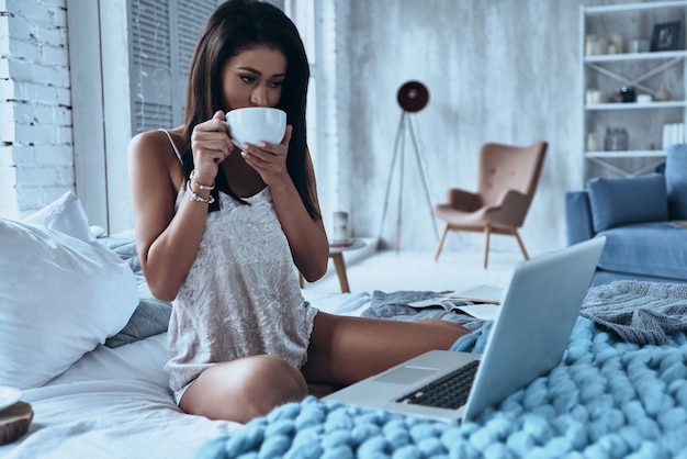 A manhã começa com café e Internet. Mulher jovem e atraente sorridente bebendo café e olhando para o laptop enquanto está sentado na cama em casa