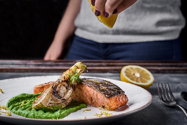 A mão aperta o limão sobre o bife de salmão assado com purê de ervilhas, cebolas caramelizadas.