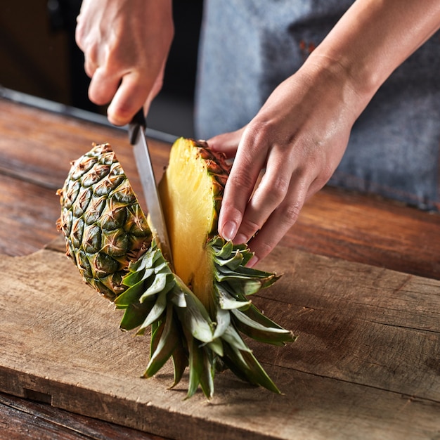 A mão da garota com uma faca corta um abacaxi de frutas tropicais em uma placa sobre uma mesa de madeira marrom em torno de um fundo escuro. Fruta saudavel