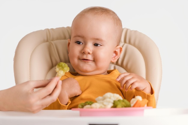 A mão da mãe dá um bebê sentado em uma cadeira de alimentação brócolis cozido