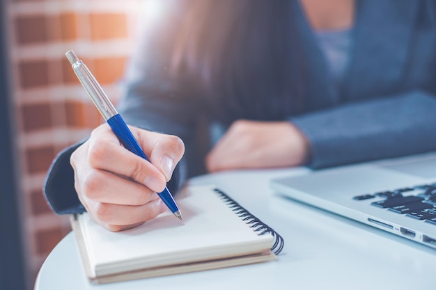 A mão da mulher está escrevendo em um bloco de notas com uma caneta e tem um computador portátil na mesa no escritório