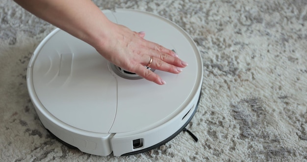 A mão da mulher pressiona o botão para ligar o aspirador robô moderno Aspirador robótico na sala de estar Limpeza doméstica