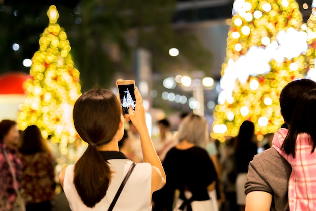 A mão da mulher que guarda o telefone celular toma uma foto da árvore e da luz de natal.