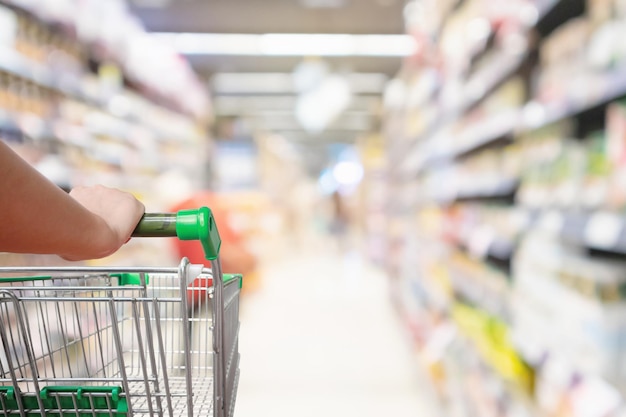 A mão da mulher segura o carrinho de compras com fundo desfocado do corredor do supermercado de desfoque abstrato