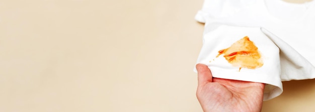 A mão da mulher segura roupas de camisa branca com mancha de ketchup em um banner de fundo bege